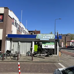 Gevel van kringloopwinkel De Nieuwe Start met blauwe luifel, fietsen geparkeerd ervoor en helder weer