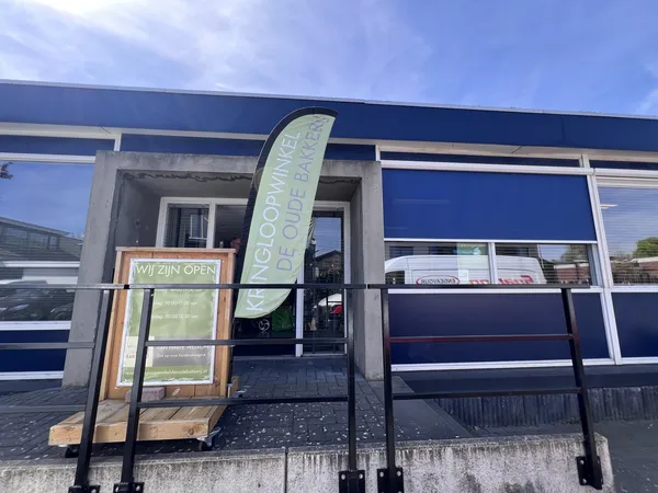 Gevel van kringloopwinkel met blauwe puien, ingang met trap en openingsbord