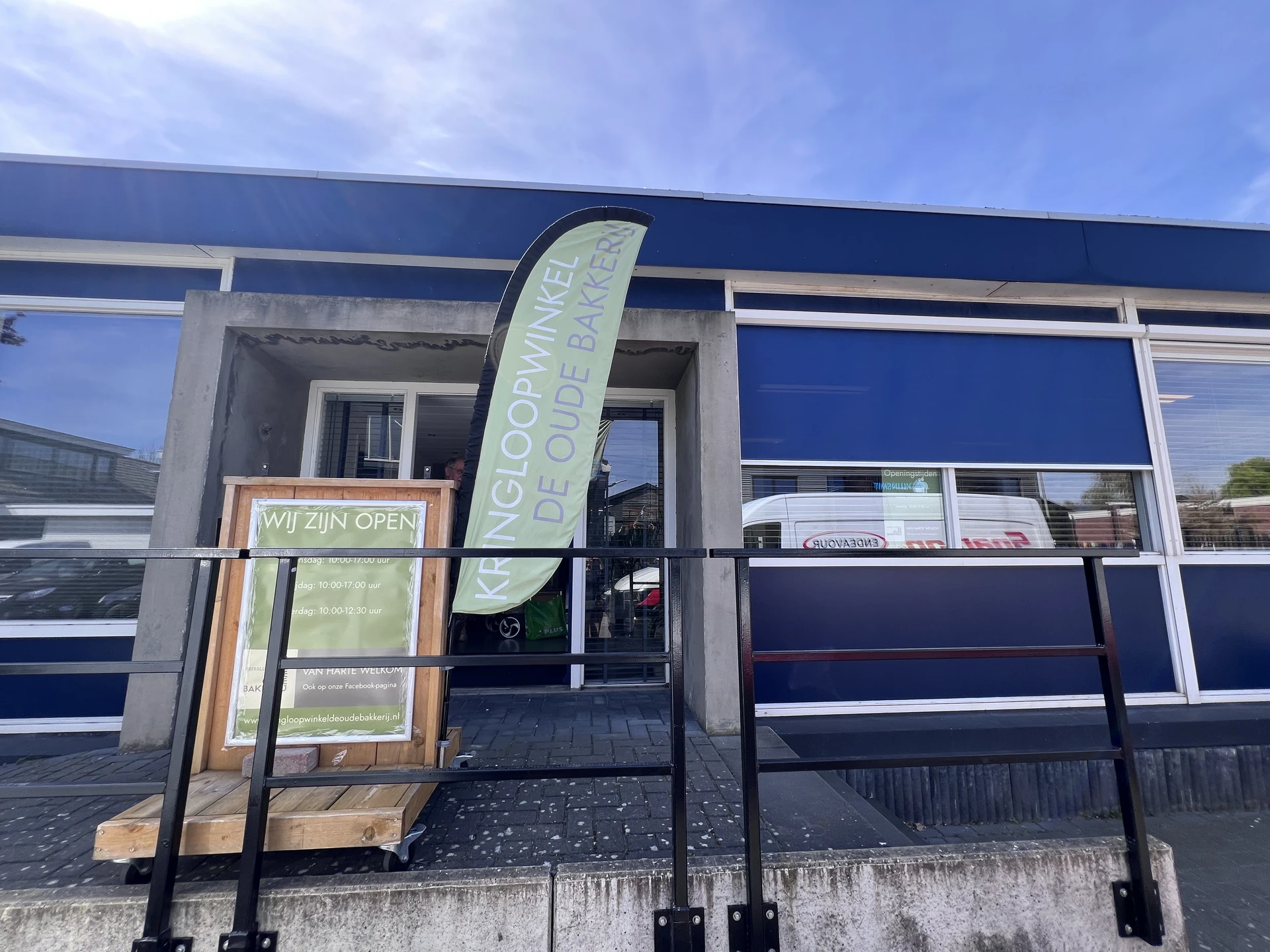 Gevel van kringloopwinkel met blauwe puien, ingang met trap en openingsbord
