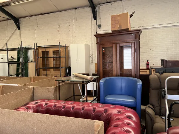 Magazijn met gestapelde dozen, houten meubels, blauwe en rode stoelen en diverse huisraad
