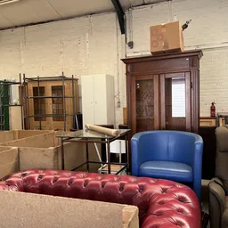 Magazijn met gestapelde dozen, houten meubels, blauwe en rode stoelen en diverse huisraad