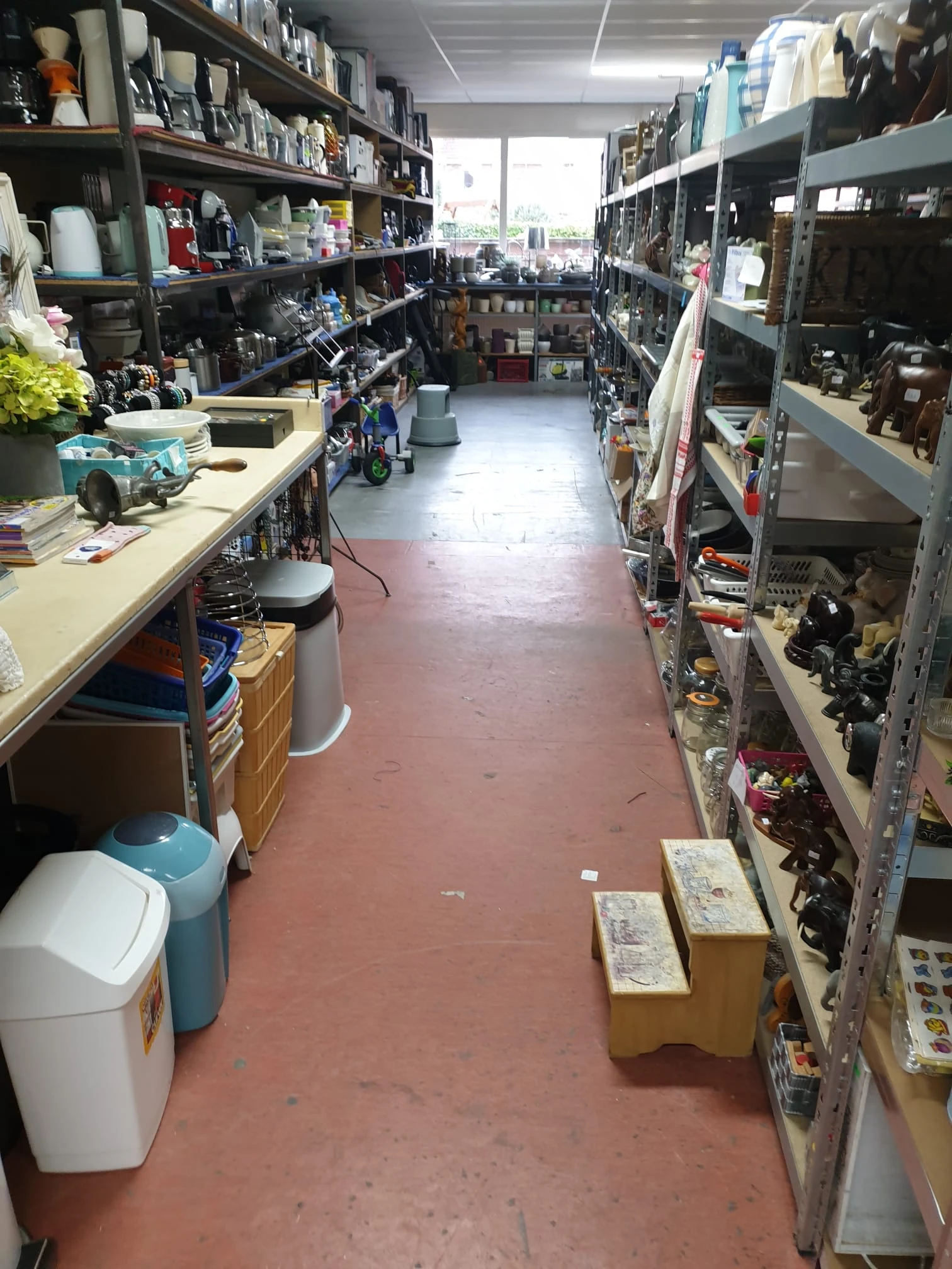 Binnenaanzicht van kringloopwinkel met rode vloer, toonbank links, stellingen met spullen rechts en diverse items uitgestald
