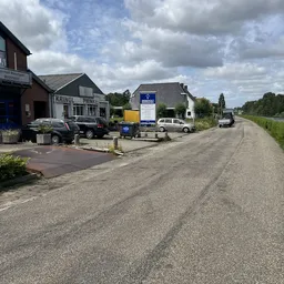 Voorgevel van kringloopwinkel met parkeerplaats, auto's en bedrijfsbussen langs een landelijke weg
