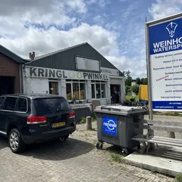 Voorkant kringloopwinkel met witte gevel, zwarte ramen en ingang, parkeerplaats met auto en afvalcontainer