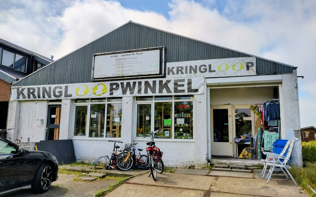 Witte gevel van kringloopwinkel met groene letters KRINGLOOOPWINKEL, fiets ervoor, parkeerplaats aan linkerkant
