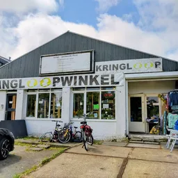 Witte gevel van kringloopwinkel met groene letters KRINGLOOOPWINKEL, fiets ervoor, parkeerplaats aan linkerkant