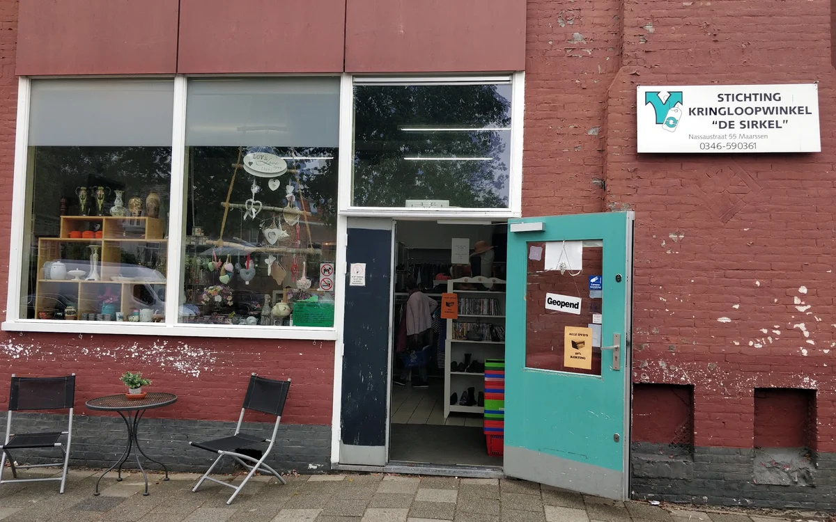 Voorgevel van kringloopwinkel met rode bakstenen muur, turquoise deur, etalageramen en twee lege stoelen op het terras