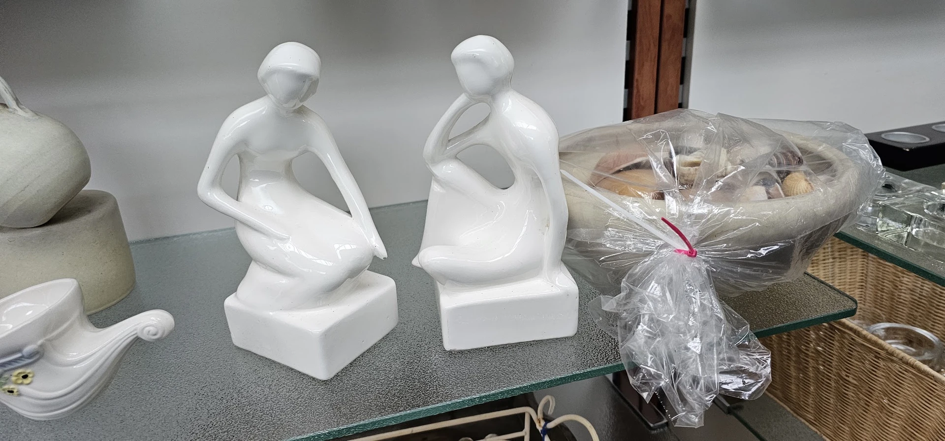 Twee witte keramische beeldjes van zittende figuren op een grijs oppervlak, naast een verpakt artikel