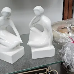 Twee witte keramische beeldjes van zittende figuren op een grijs oppervlak, naast een verpakt artikel