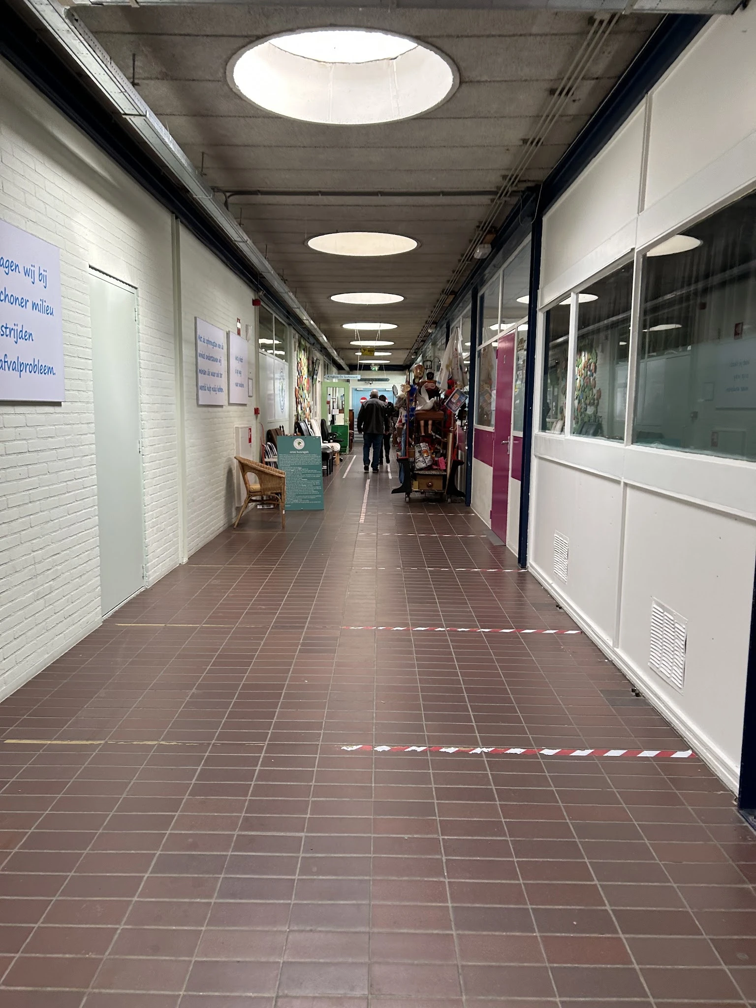 Winkelgang met witte en bordeauxrode deuren, wit getegelde muren, verlicht plafond en voetverkeer in de verte