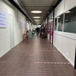 Winkelgang met witte en bordeauxrode deuren, wit getegelde muren, verlicht plafond en voetverkeer in de verte