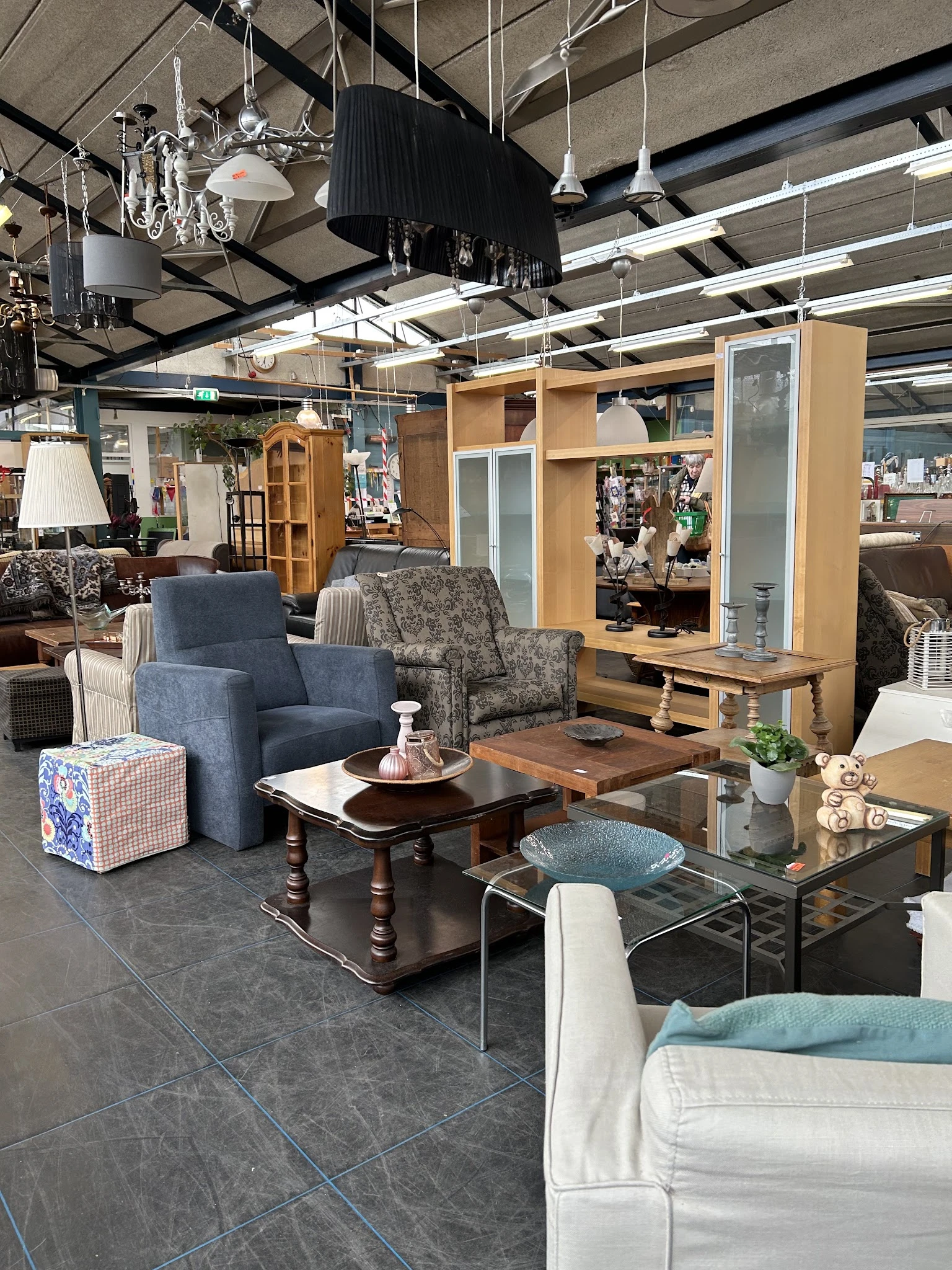 Winkelvloer met diverse meubels: blauwe fauteuil, gebloemde stoel, houten kasten en tafels, speelgoed, plantjes en lampen.