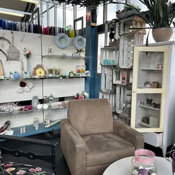 Binnenkant van kringloopwinkel met grijze fauteuil, witte stellingen met decoratie-items, kasten en hangend huisraad