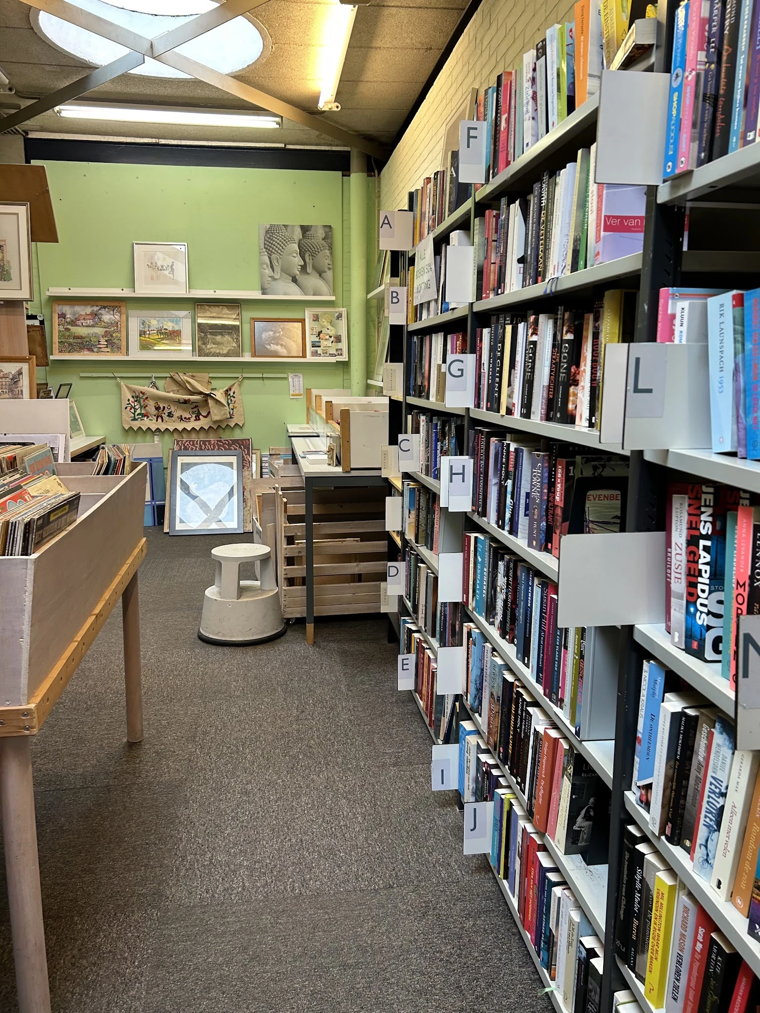 Winkelinterieur met boekenrekken, groene muur met kunstwerken, toonbank en witte poef op grijze vloer