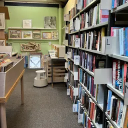 Winkelinterieur met boekenrekken, groene muur met kunstwerken, toonbank en witte poef op grijze vloer