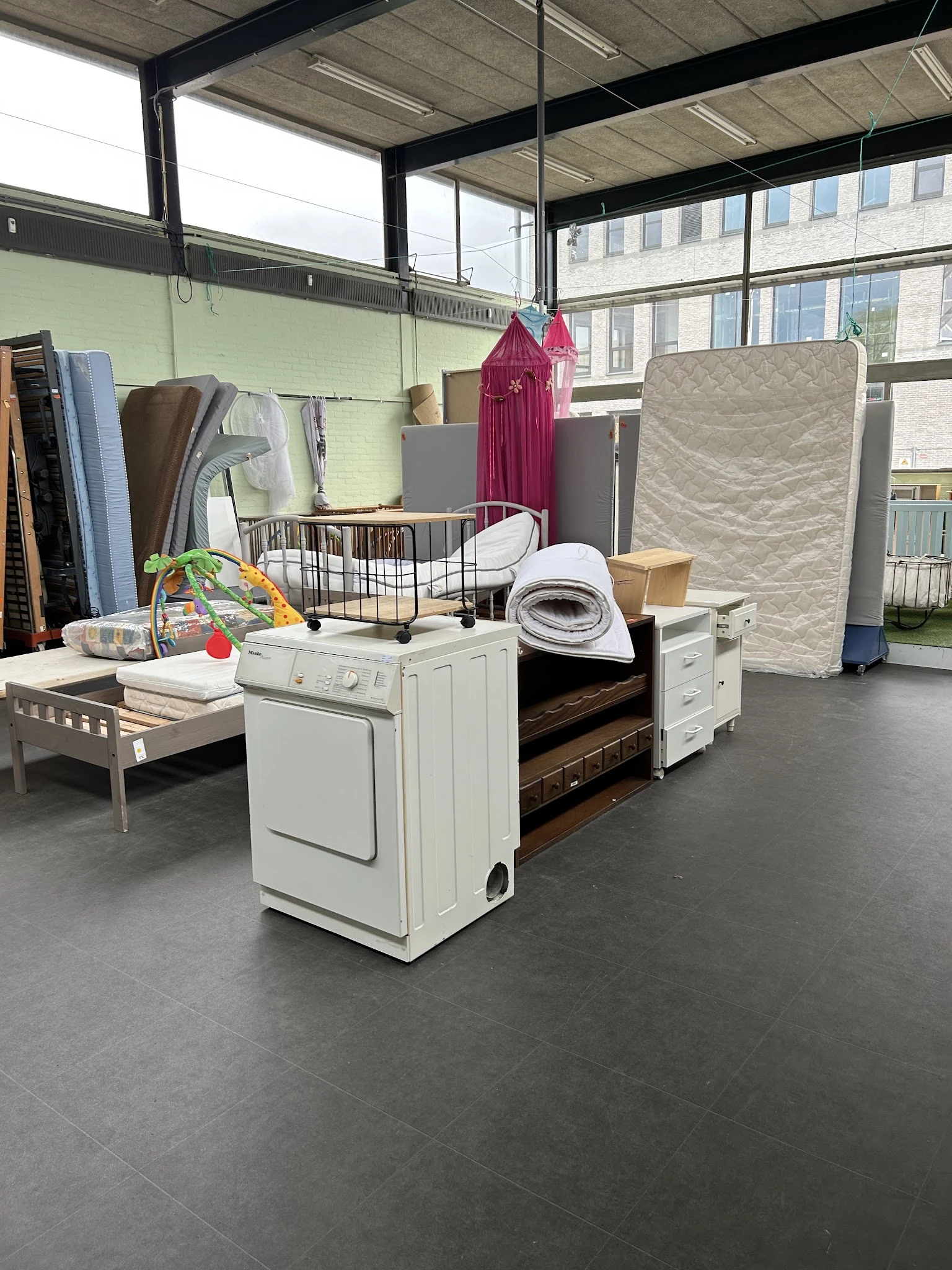 Uitgestalde meubels en huishoudartikelen in kringloopwinkel, waaronder bedframe, wasdroger, matrassen en textiel