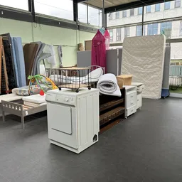 Uitgestalde meubels en huishoudartikelen in kringloopwinkel, waaronder bedframe, wasdroger, matrassen en textiel