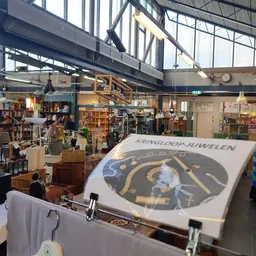 Ruim interieur van een kringloopwinkel met diverse afdeling, stellingen vol spullen en een groot logo-banner van Kringloop Ju