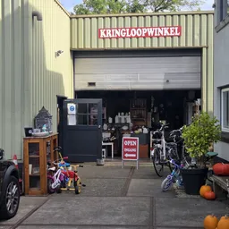 Voorgevel van kringloopwinkel met open garagedeur, uithangbord 'KRINGLOOPWINKEL', fietsen en speelgoed buiten
