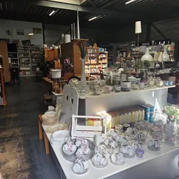 Overzicht van kringloopwinkel met toonbanken vol glaswerk, servies en huishoudartikelen, stellingen op achtergrond