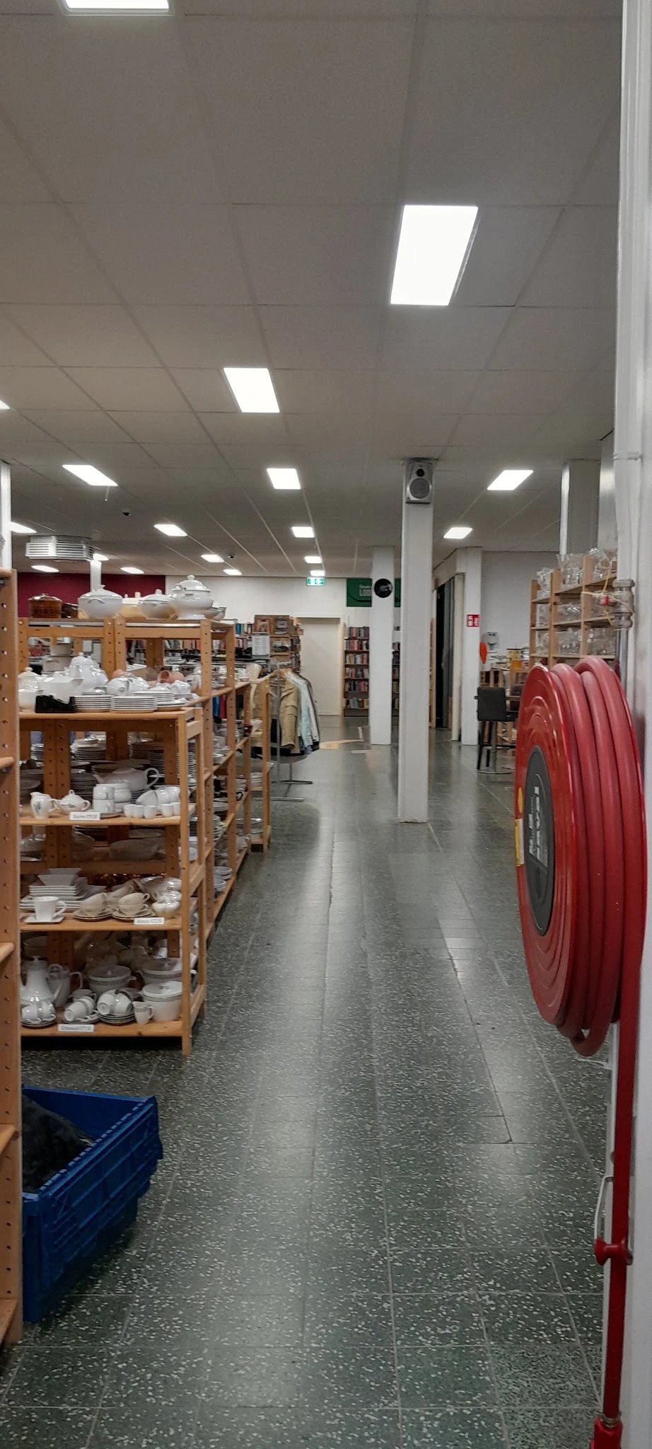 Winkelinterieur met houten stellingen vol servies, kleding aan rekken, boekenkasten en een rode brand-slang aan de rechterkan