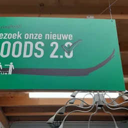 Groen bord met witte tekst 'Bezoek onze nieuwe LOODS 2.5' en afstandssymbool, hangend aan plafond