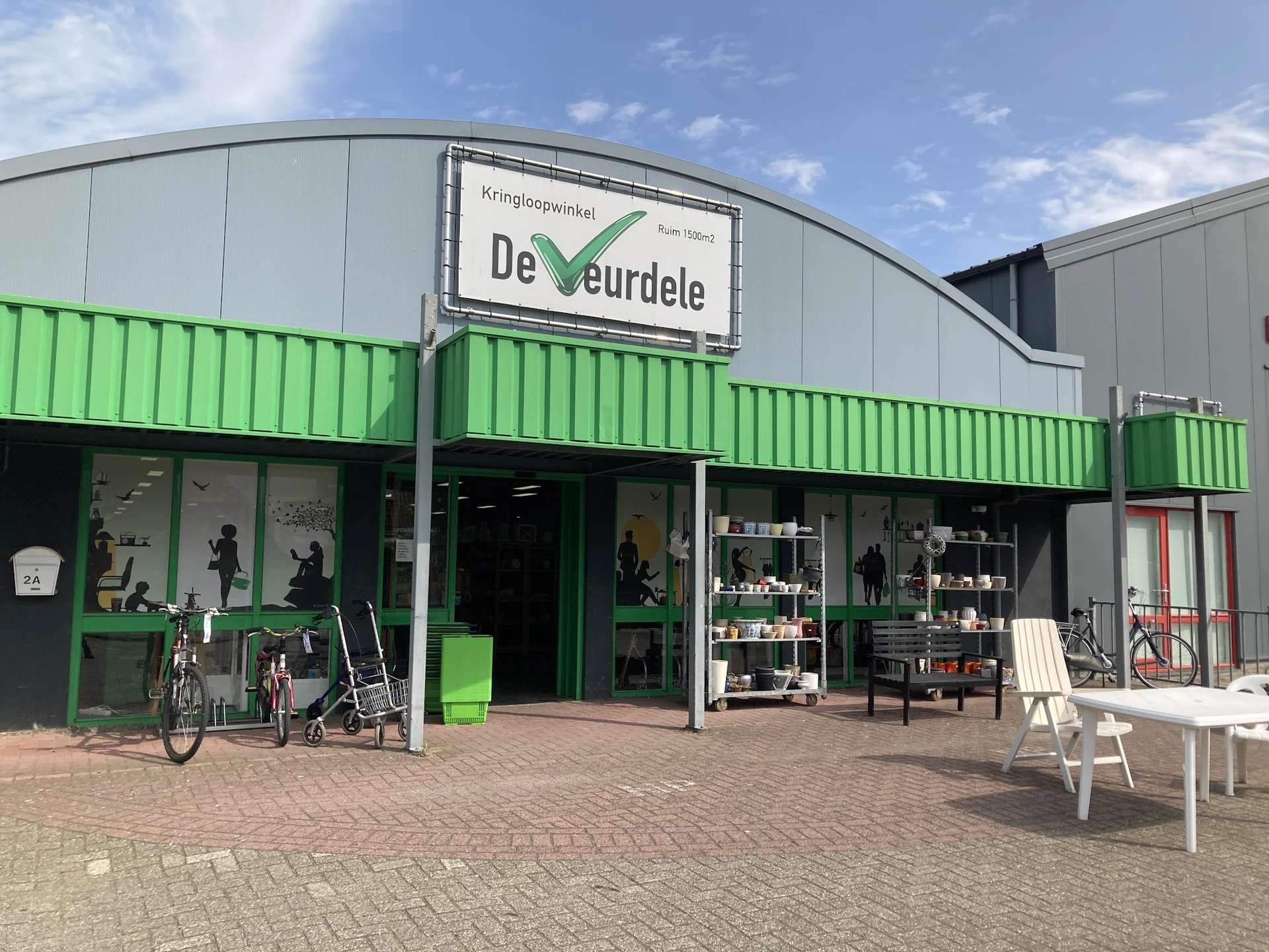 Gevel van kringloopwinkel De Veurdele met groene metalen wanden, etalageramen en ingang aan Hellingstraat