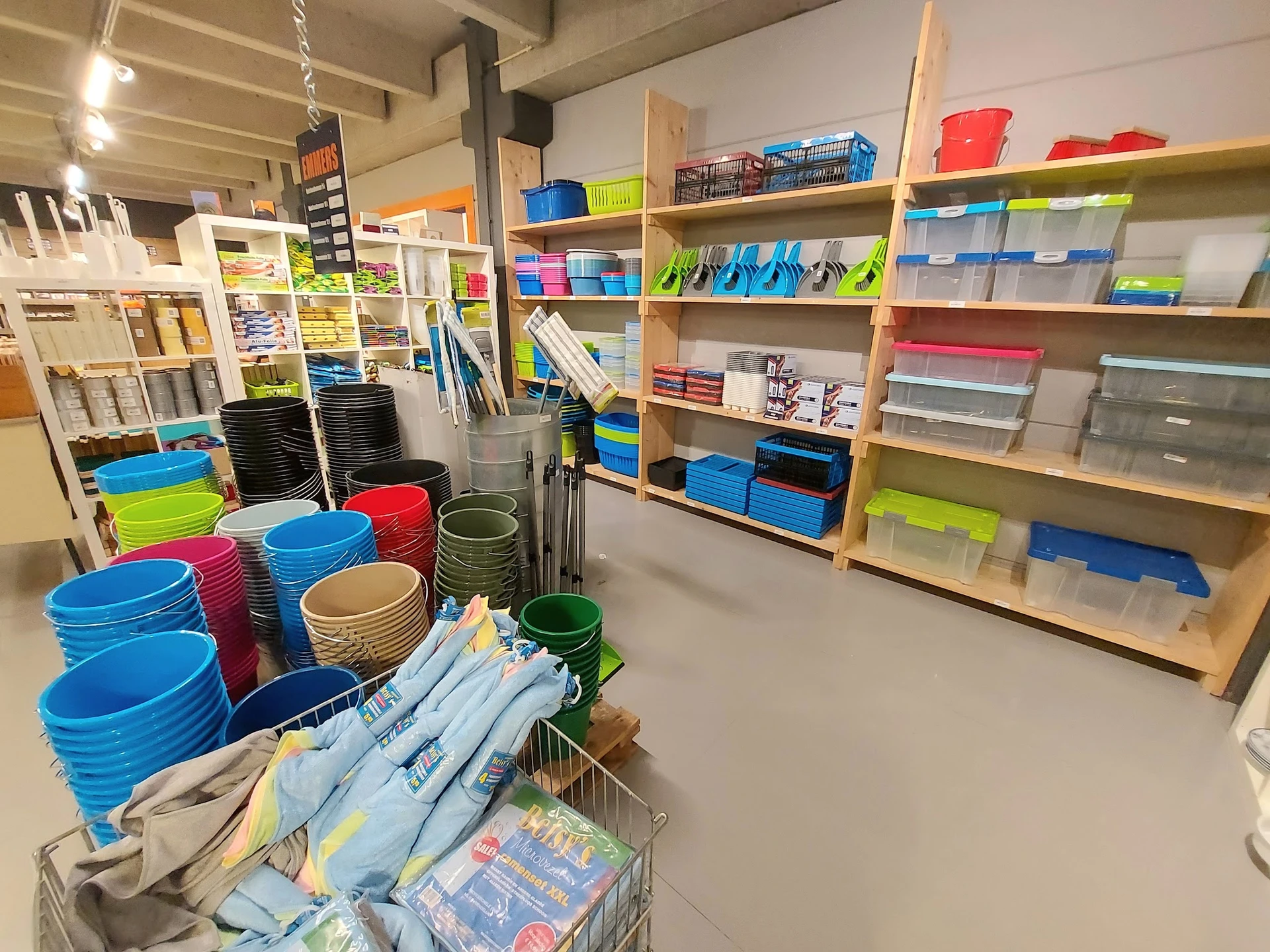 Winkelrek met plastic bakken en opbergdozen in verschillende kleuren, met winkelwagen op voorgrond