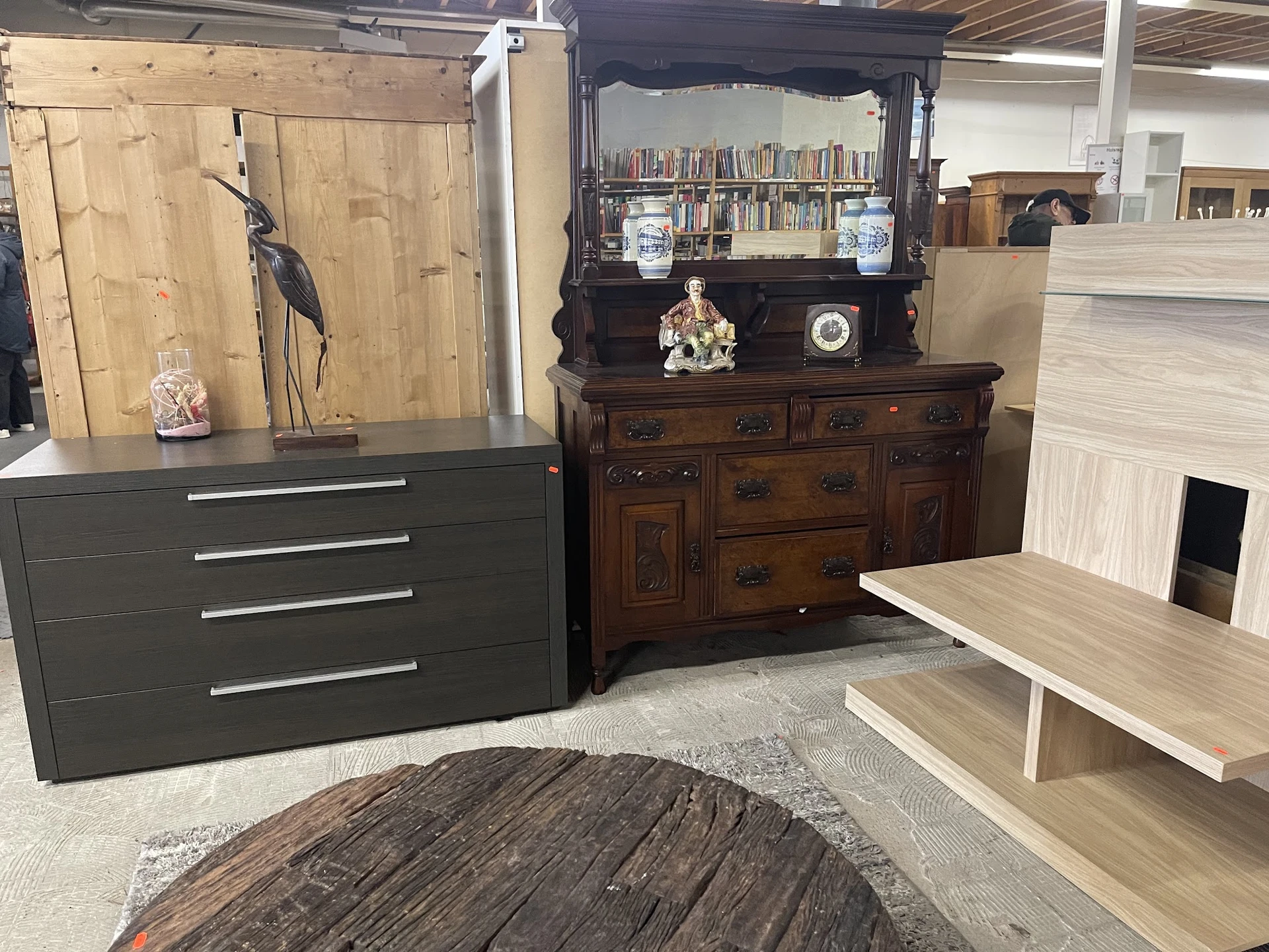 Showroom met diverse meubelen: zwarte ladekast, donkerbruin dressoir met boekenkast, houten tafel en decoratieve objecten op 