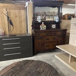 Showroom met diverse meubelen: zwarte ladekast, donkerbruin dressoir met boekenkast, houten tafel en decoratieve objecten op 