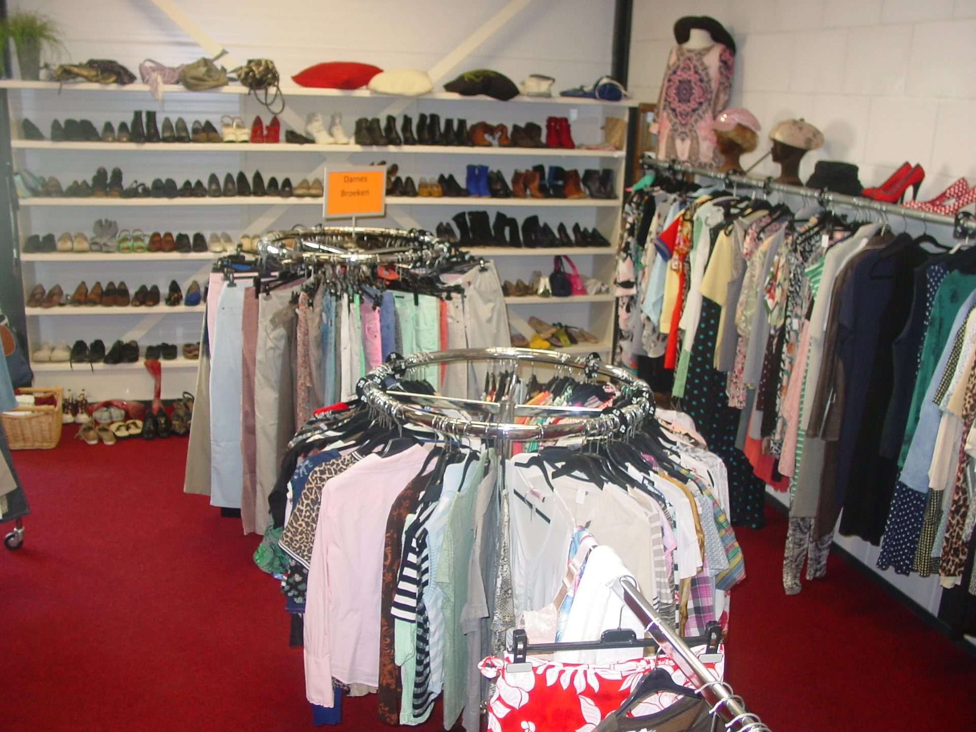 Rekken met tweedehands kleding, schoenen op planken achter, rode vloer in kringloopwinkel