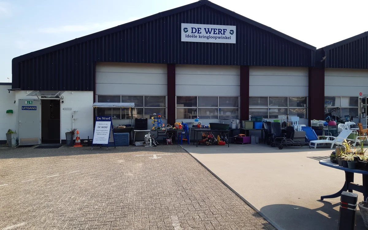 Voorgevel van kringloopwinkel De Werf met zwart dak, witte garagedeuran en parkeerplaats met tweedehandsartikelen