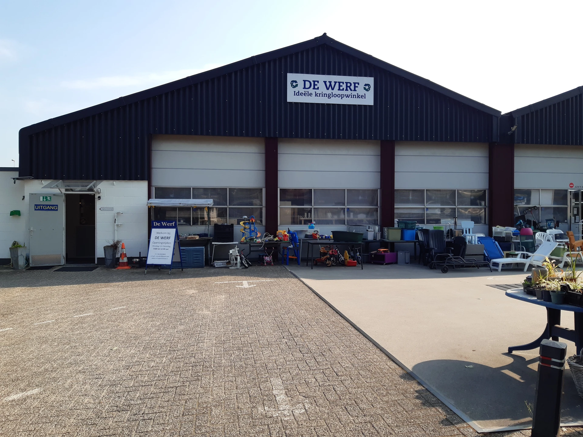 Voorgevel van kringloopwinkel De Werf met zwart dak, witte garagedeuran en parkeerplaats met tweedehandsartikelen