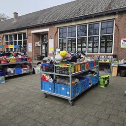 Uitgestalde tweedehands spullen op karren en tafels buiten de kringloopwinkel, inclusief huishoudartikelen, speelgoed en dive