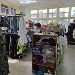Kringloopwinkel met kledingrekken, houten kastje met hoeden en accessoires, grote ramen met uitzicht