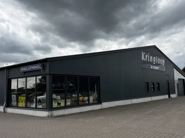 Zwart pand van kringloopwinkel met naam 'Kringloop' op gevel, grote ramen aan voorkant, bakstenen plein ervoor