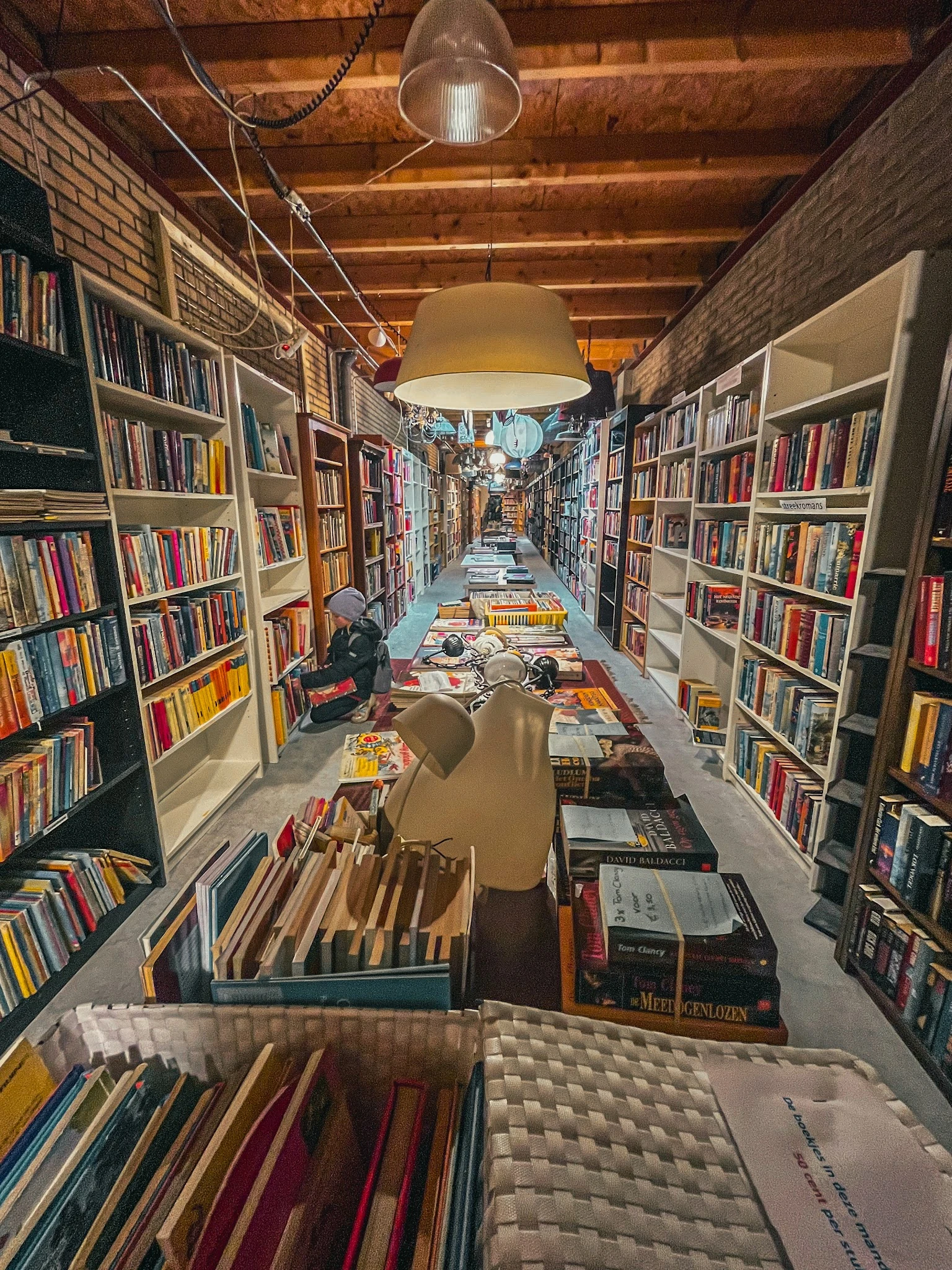 Lange smalle winkelgang met boekenkasten aan beide zijden, boeken op tafels in het midden, hangende lamp, zicht naar het acht
