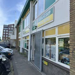Gevel van kringloopwinkel met witte en groene afwerking, gesloten met rolgordijn, gele raamkozijnen en groene winkelnaamborde