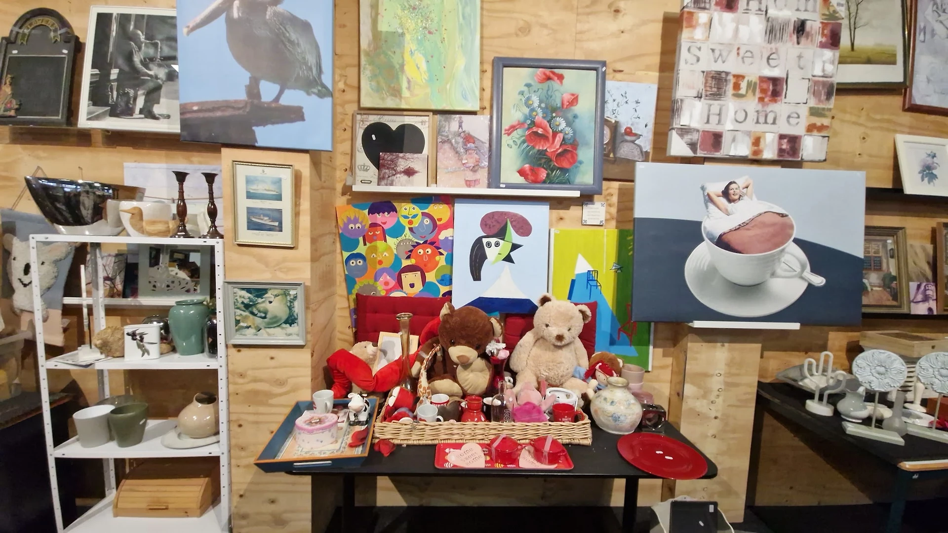 Uitgestalde tweedehandsartikelen op een tafel: knuffels, servies, kunstwerken en decoratieve items tegen een houten achtergro