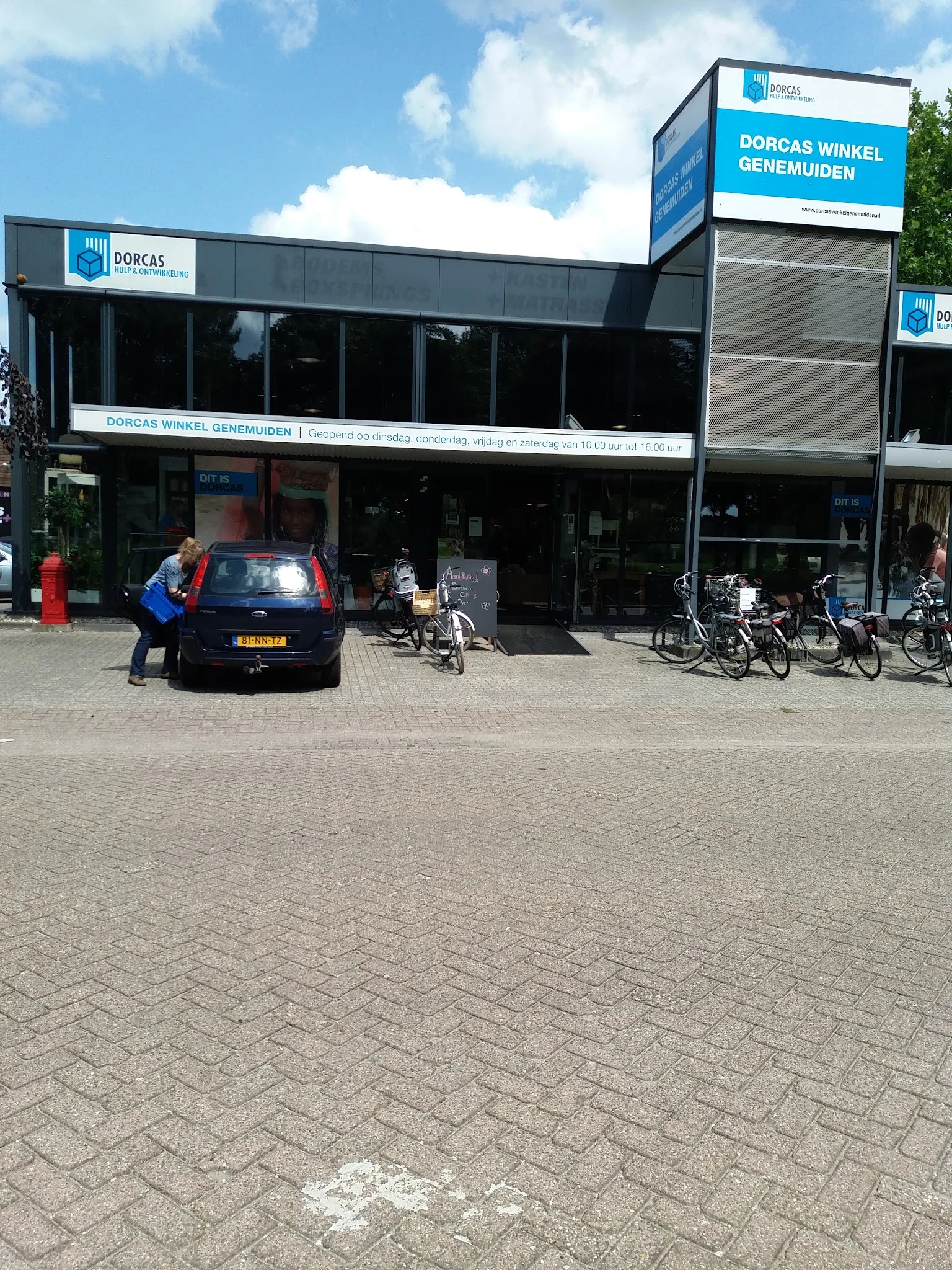 Voorgevel van kringloopwinkel met donkere etalage, fietsen ernaast geparkeerd en man in blauwe jas