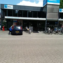 Voorgevel van kringloopwinkel met donkere etalage, fietsen ernaast geparkeerd en man in blauwe jas