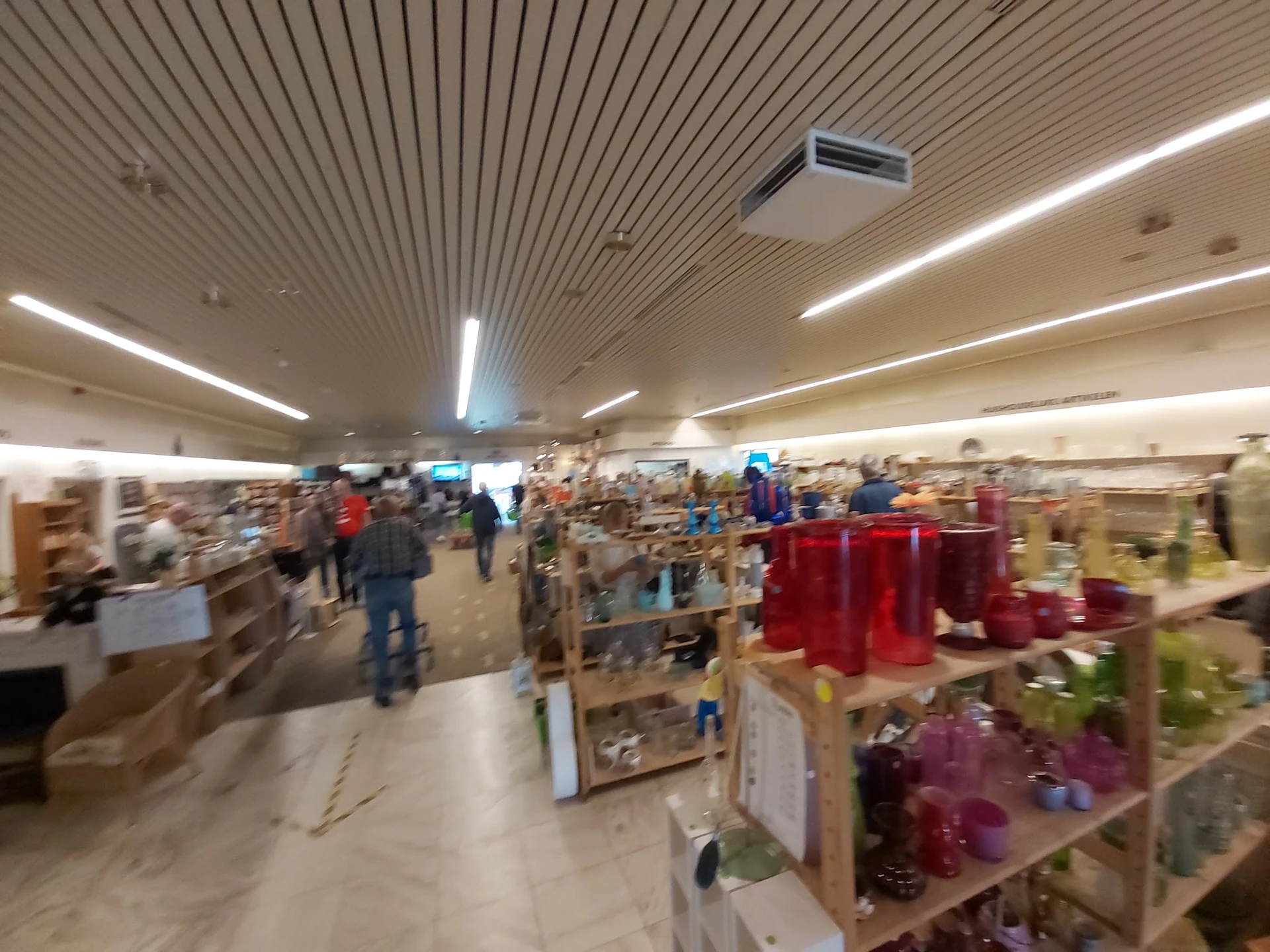 Overzicht van de winkelvloer met verschillende stellingen, artikelen en bezoekers in de kringloopwinkel