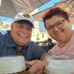 Twee personen zitten samen onder een parasol met drankjes, met een gebouw op de achtergrond