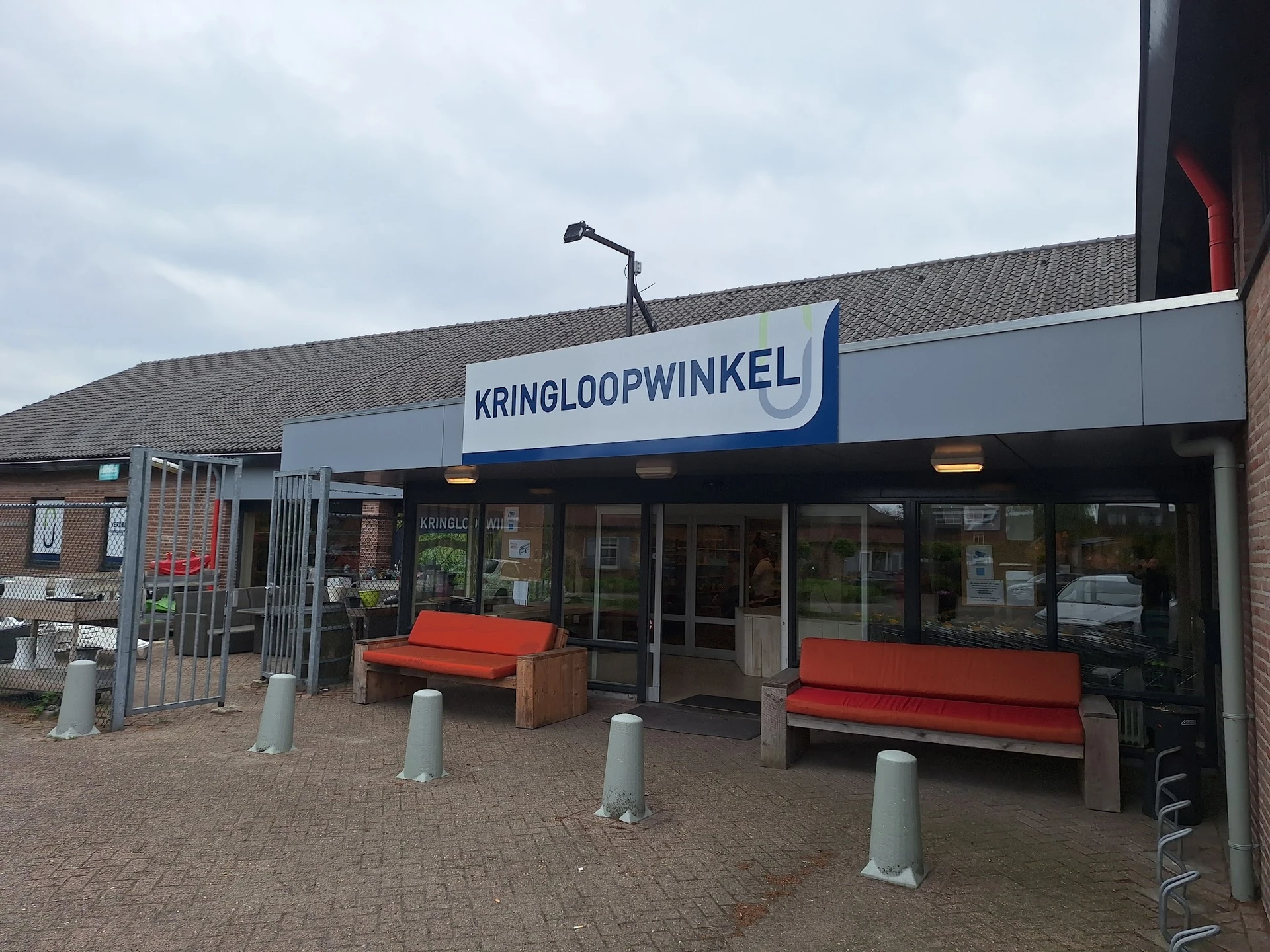 Winkelgevel met blauwe zonnescherm waarop 'KRINGLOOPWINKEL' staat, rode zitbanken ervoor en betonnen zuilen op tegelverhardin