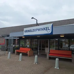 Winkelgevel met blauwe zonnescherm waarop 'KRINGLOOPWINKEL' staat, rode zitbanken ervoor en betonnen zuilen op tegelverhardin