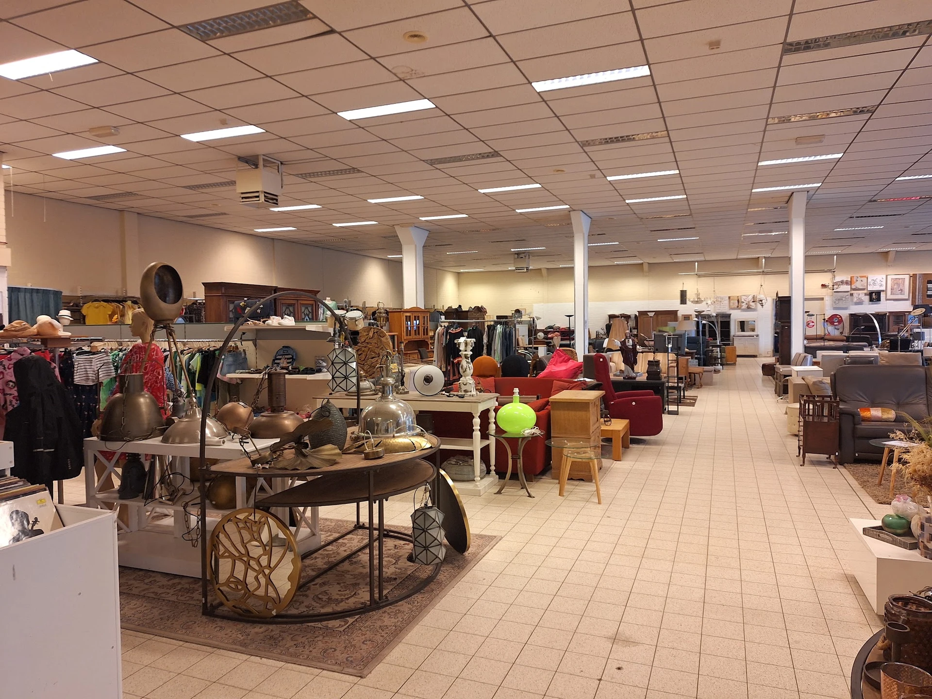 Ruim winkelinterieur met diverse tweedehands meubels, kleding en huishoudelijke artikelen uitgestald op de vloer en aan rekke