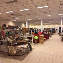 Ruim winkelinterieur met diverse tweedehands meubels, kleding en huishoudelijke artikelen uitgestald op de vloer en aan rekke
