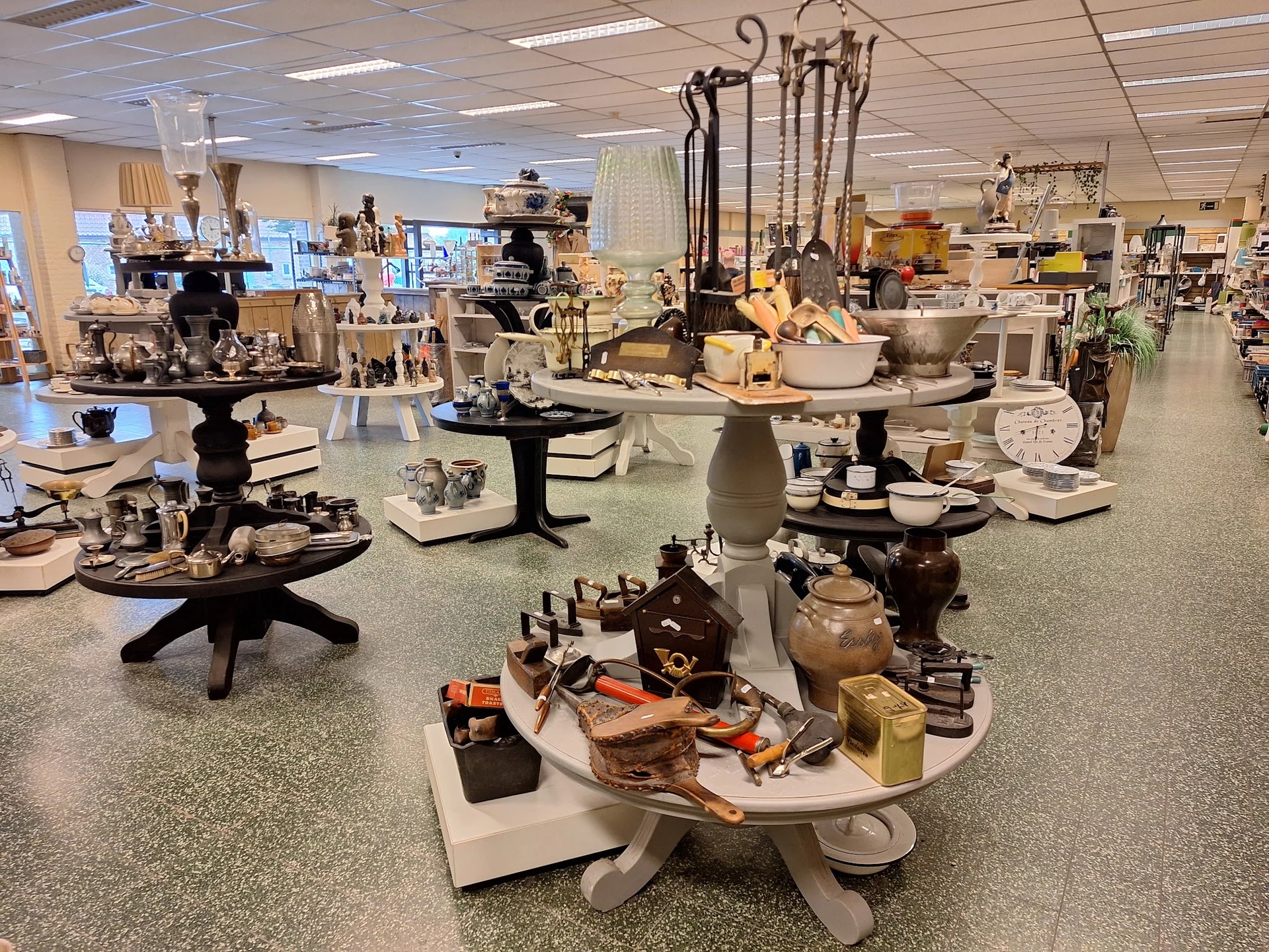 Diverse huishoudelijke artikelen en decoratieve items uitgestald op tafels in de winkel, waaronder servies, bekers, kandelaar
