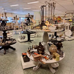 Diverse huishoudelijke artikelen en decoratieve items uitgestald op tafels in de winkel, waaronder servies, bekers, kandelaar