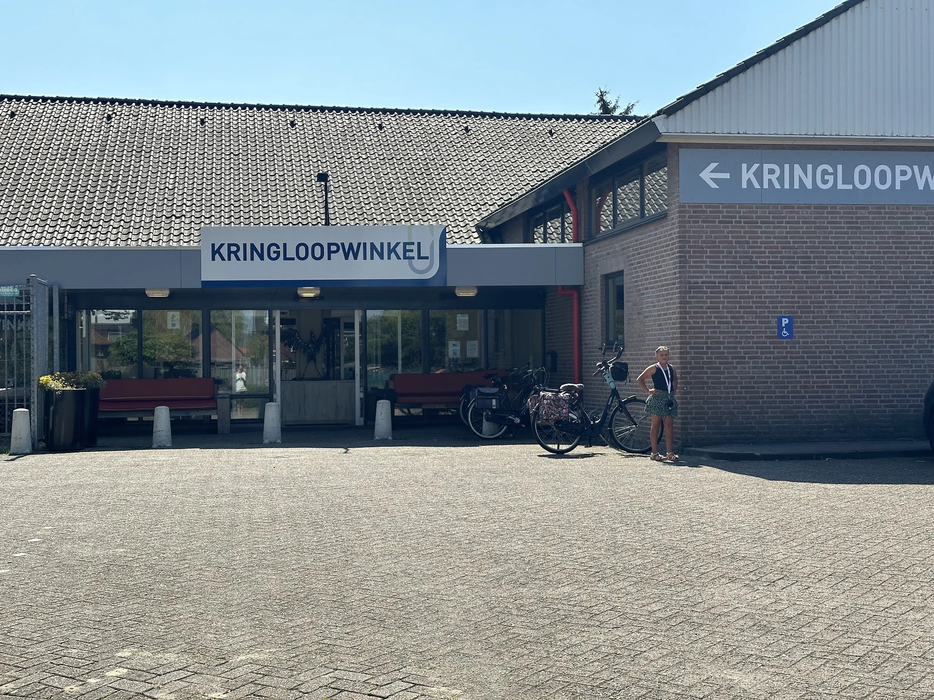 Voorgevel van kringloopwinkel met blauwe luifel en winkelnaam, fiets geparkeerd ervoor